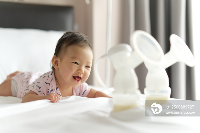 Breast milk in milk pump’s bottles on the bed with selective focus on smiling crawling baby. The milk got from milk pump’s machine and ready for the baby. Baby health care concept.