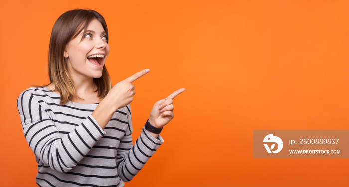 Portrait of surprised happy young woman with brown hair in striped shirt standing, pointing at empty space for announcement, unbelievable news. indoor studio shot isolated on orange background