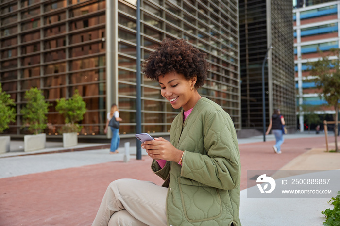 Sideways shot of beautiful woman with Afro hair holds smartphone reads received sms message has cheerful expression sits in downtown against cityscrapers dials number of taxi service. Technology