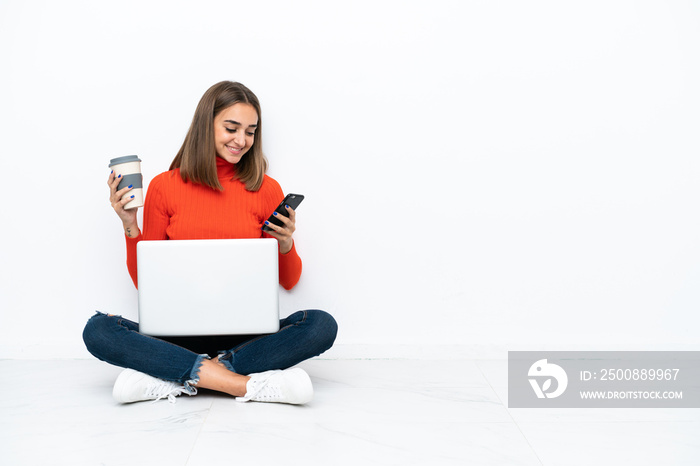 Young caucasian woman sitting on the floor with a laptop holding coffee to take away and a mobile