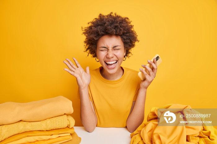 Cheerful dark skinned woman with curly hair gets excellent news holds mobile phone raises palm feels very happy sits at table near pile of unfolded laundry stack of folded clothes yellow background