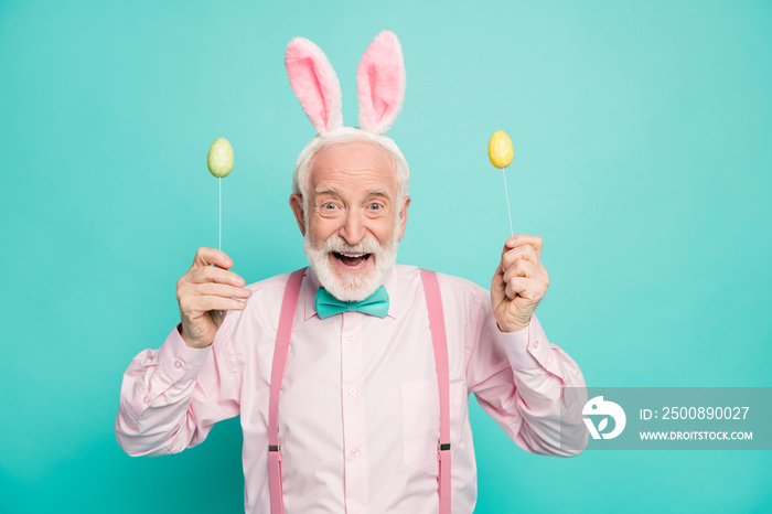 Portrait of astonished funky crazy excited old man hold stick with colorful eggs enjoy easter party event wear pink hare headband pink shirt isolated over turquoise color background