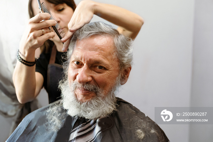 Working with hair scissors. Professional hairstyler woman cutting hair of mature man. He is getting haircut at barbershop or beauty salon.