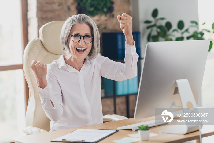 Photo of smiling lucky lady boss wear white shirt eyeglasses rising fists successful startup indoors workplace workstation