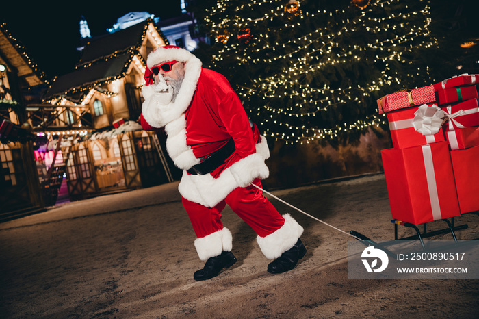 Full body portrait of aged man santa claus costume step walk pull sled pile stack box package show shh gesture magic tree garland lights outside