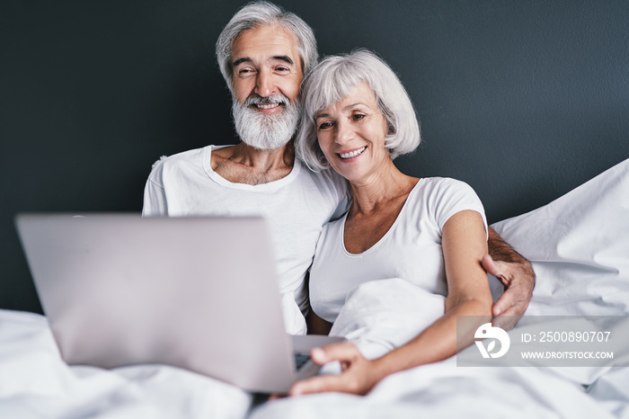 Happy family. Cozy home. Beautiful senior woman and her husband using laptop computer together in bedroom.