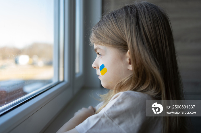 A little girl looking in the window of her house with a painted yellow-blue flag of Ukraine, stop the war, protest and patriot, hope for peace.