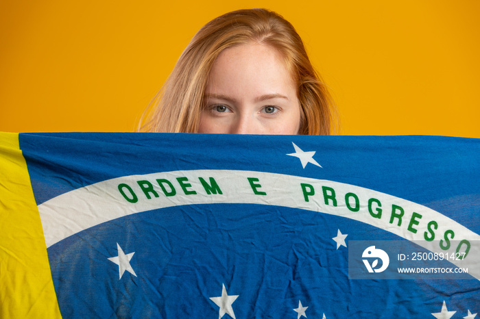 Mysterious redhead woman fan holding a Brazilian flag in your face. Brazil colors in background, green, blue and yellow. Elections, soccer or politics.