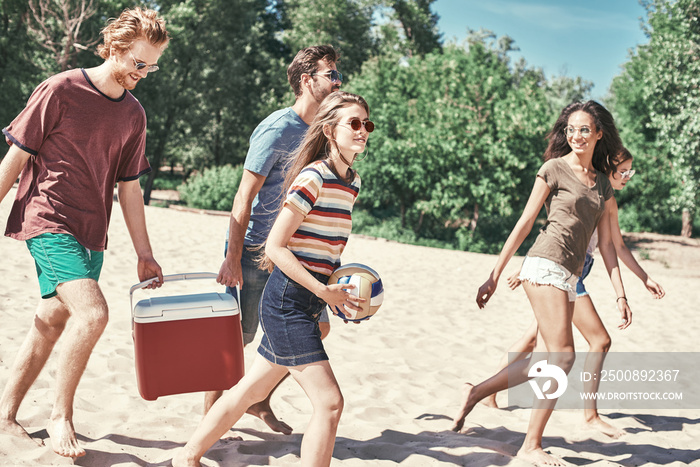 Friends on the beach. Rear view of cheerful young people walking by the beach to the sea while two men carrying plastic cooler