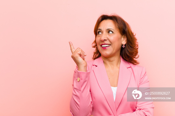 middle age woman smiling happily and looking sideways, wondering, thinking or having an idea against pink wall