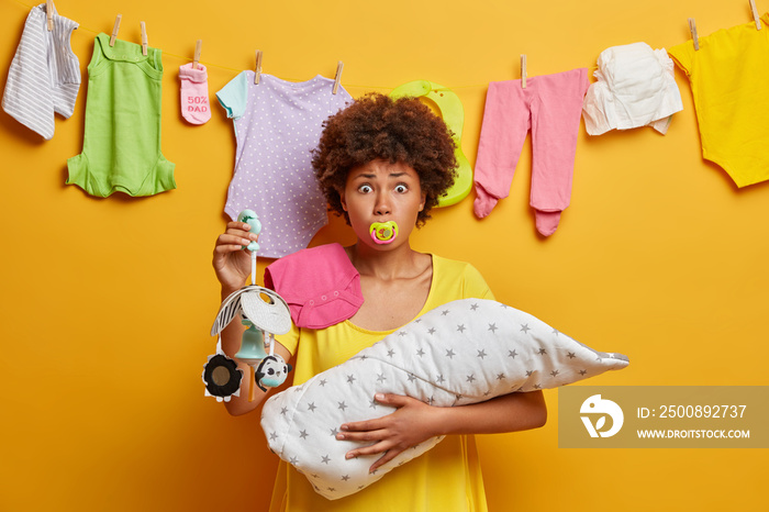 Scared young mother poses with nipple in mouth, holds mobile in hand, cares about newborn child, busy nursing baby, soothes crying daughter, shows toy for crib. Family and babysitting concept