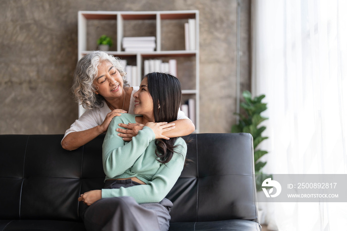 Loving adult daughter hugging older mother, standing behind couch at home, family enjoying tender moment together, young woman and mature mum or grandmother looking at each other, two generations