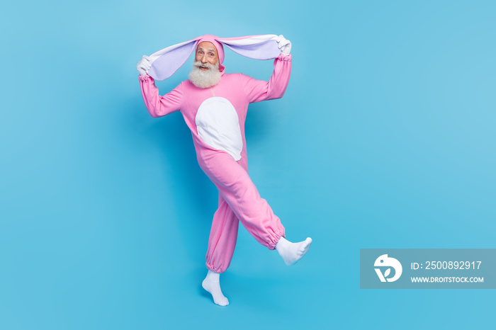 Full length photo of funky carefree retired guy wear pink hare sleepwear walking holding ears isolated blue color background