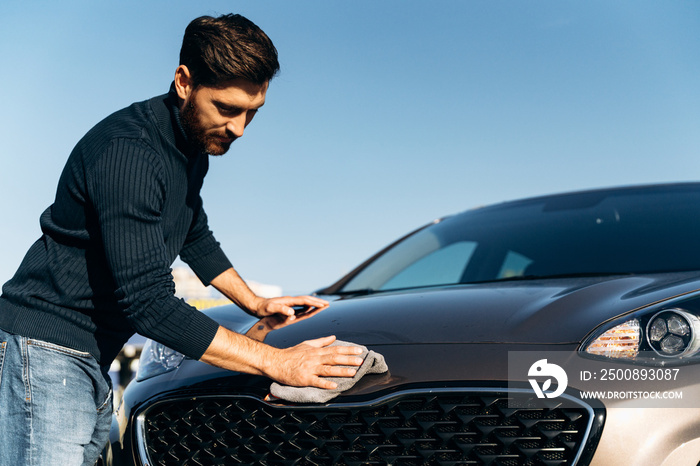 Low angle shot of a happy young man polishing his car with microfiber cloth while feeling satisfied during the sunny summer day. Stock photo