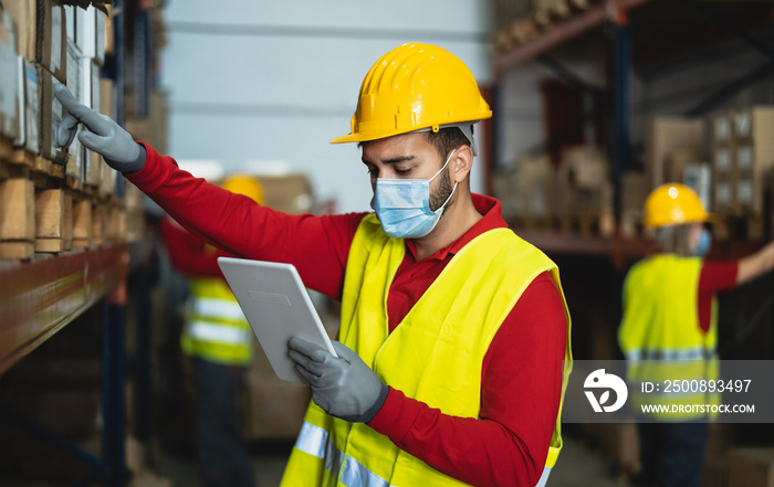 Young man working in warehouse doing inventory using digital tablet and loading delivery boxes plan while wearing face mask during corona virus pandemic - Logistic and industry concept