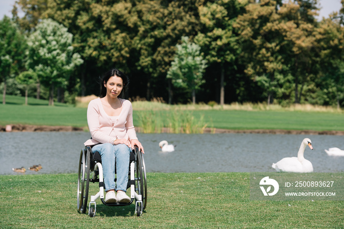 Young disabled woman looking at camera while sitting in wheelchair near pond with white swans