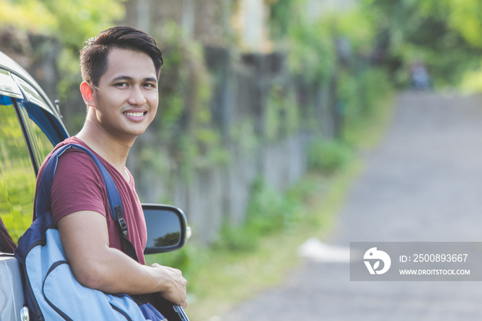 asian male student with backpack smiling leaning on his car