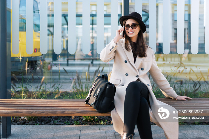 Casual woman in glasses sitting at the bus stop and talking on the mobile phone. Passenger waiting for city bus