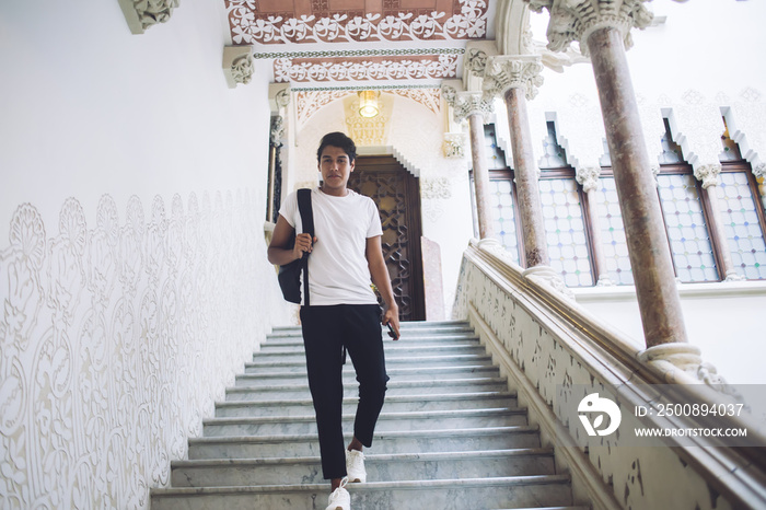 Full length portrait of handsome Latino student with backpack walking at university stairs and looking at camera, young male tourist 20 years old visiting architecture buildings during sightseeing