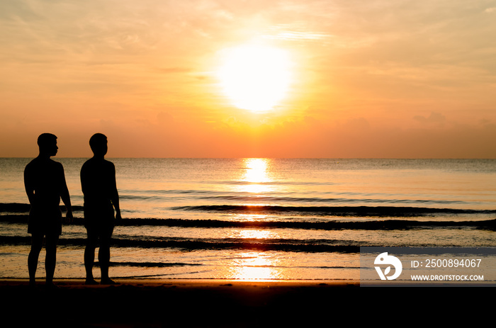 The silhouette photo of the gay couple standing together on the beach enjoy sunrise moment with the reflection on the sea in summer season for vacation trip.