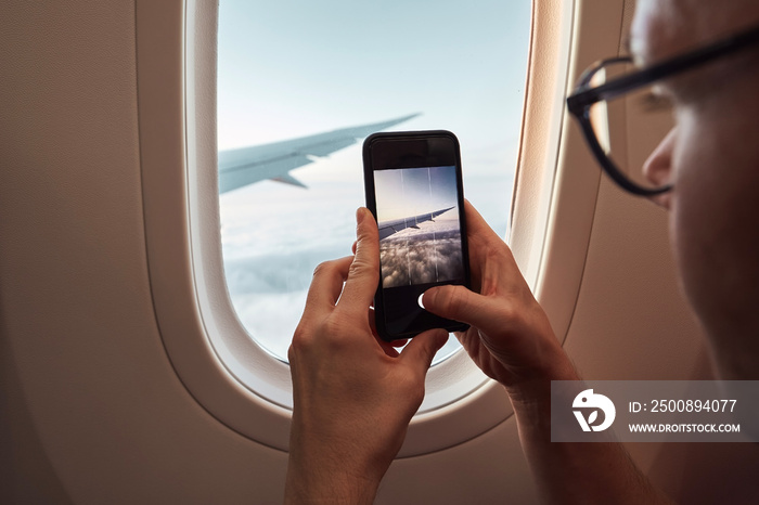 Passenger photographing through airplane window. Man using smart phone during flight above cloouds. Themes travel, connection and memories..