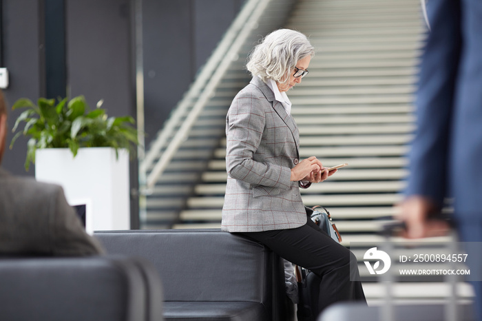 Serious senior Asian businesswoman in formalwear sitting on back of sofa in lobby and surfing internet on phone while waiting for check-in time