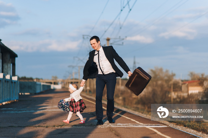 Loving dad and businessman in suit and glasses with a suitcase hurrying to the train, walking with a child and a cute daughter against the blue sky, on the platform. Travel and vacation.
