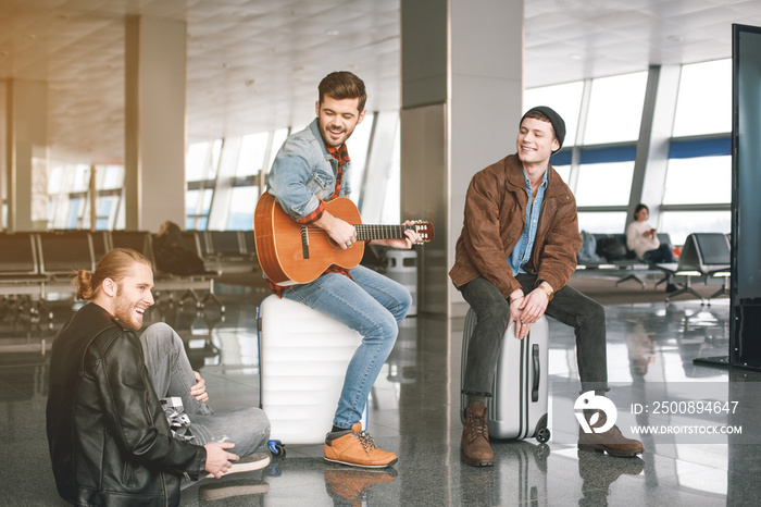 Cheerful male playing musical instrument near friends with luggages in airport. Expectation concept