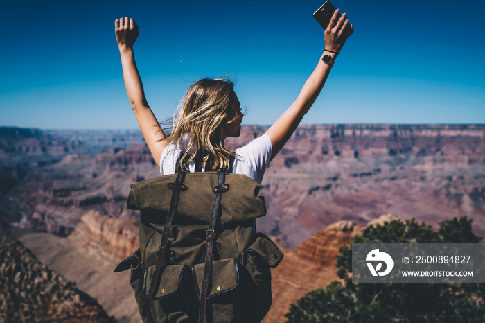 Back view of excited hipster girl standing on high mountain top feeling free raising hands up, happy female traveler with backpack celebrating achievement of reaching peak during hiking tour in USA