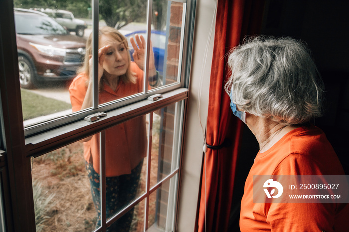 A baby boomer daughter visiting her elderly shut-in mother in her 80’s during quarantine from COVID-19 coronavirus through a window as to not catch this contagious disease