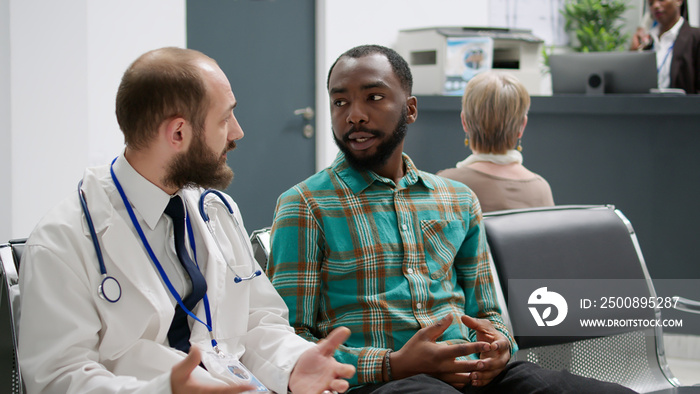 Doctor talking to african american man in hospital reception, doing checkup examination in waiting area lobby. Diverse people chatting about healthcare treatment and diagnosis at facility.