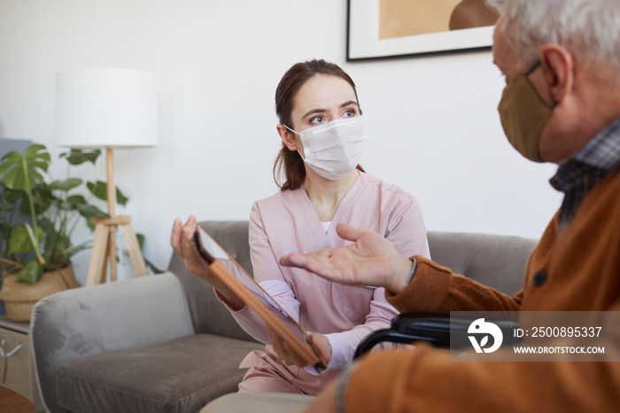 Portrait of young female nurse assisting senior man in wheelchair using digital tablet at retirement home, both wearing masks, copy space