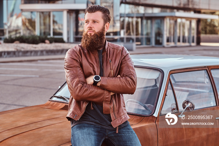 Bearded male dressed in brown leather jacket leaning on tuned retro car in the city parking.