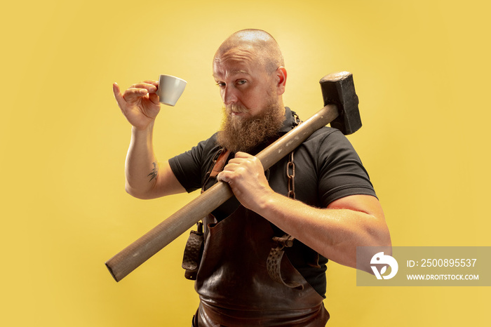 Comic portrait of muscular bearded bald man, blacksmith in leather apron or uniform isolated on yellow studio background. Concept of labor, retro professions, power, strength, humor