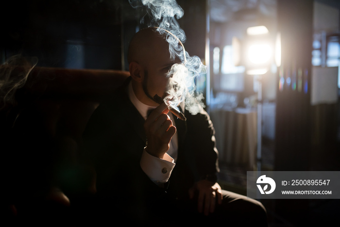 A brutal man in a dress coat smokes a cigar.