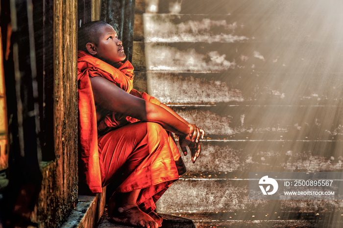 A Thai Buddhist novice sitting at old temple door close to stair steps looking up sky watching sun light