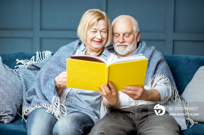 Lovely senior couple reading book while sitting wrapped with warm plaid on the couch at home