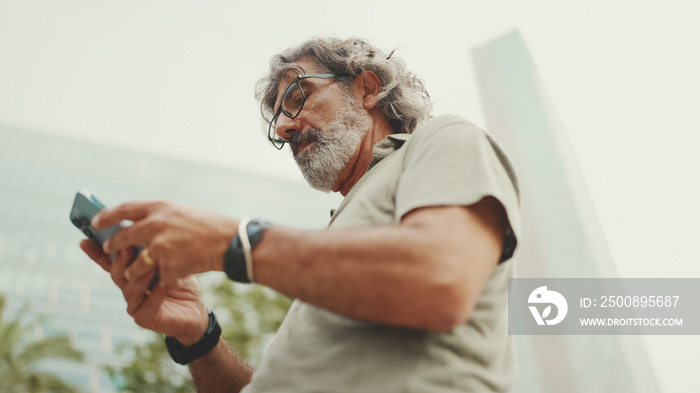 Friendly middle aged man with gray hair and beard looking at map trying to find his way using his mobile phone. Mature gentleman in eyeglasses using map app in cellphone outdoors