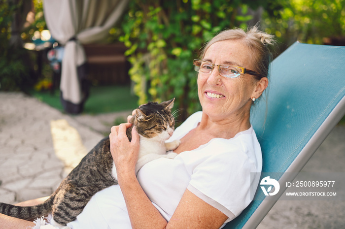 Happy smiling senior elderly woman in glasses relaxing in summer garden outdoors hugging domestic tabby cat. Retired old people and animals pets concept