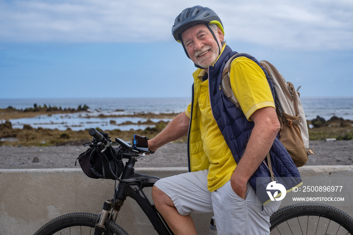 Smiling bearded cyclist senior man along the sea beach carrying a backpack, wearing helmet running with electro bicycle looking at camera. Concept of healthy and sustainable mobility