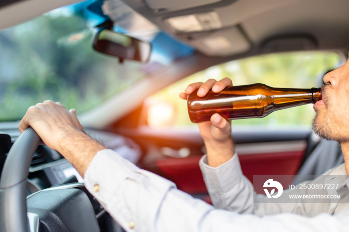 Drunk asian young man drives a car with a bottle of beer with sunset background