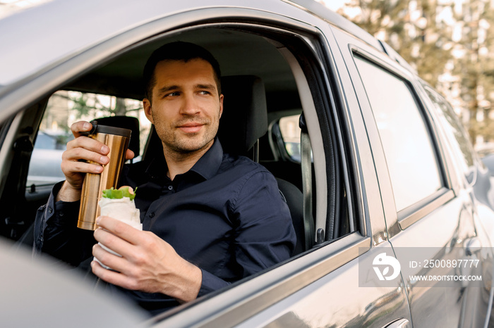 Stopping for a bite to eat . Man eat snack in the car and drinks coffee or tea.