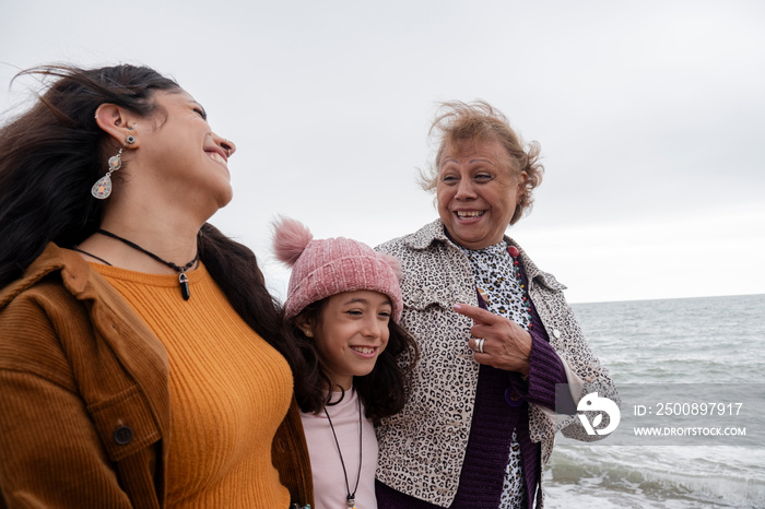 Grandmother, mother and daughter laughing while walking on beach on cloudy day