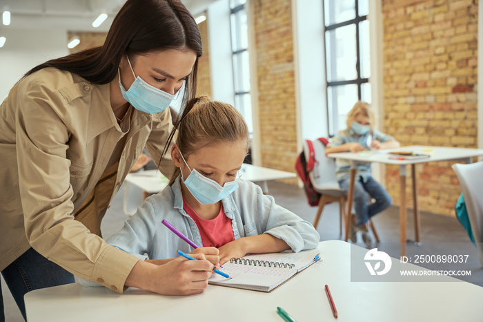 Helpful young female teacher wearing protective face mask helping little girl doing sums. Kids studying in elementary school, sitting at the desk