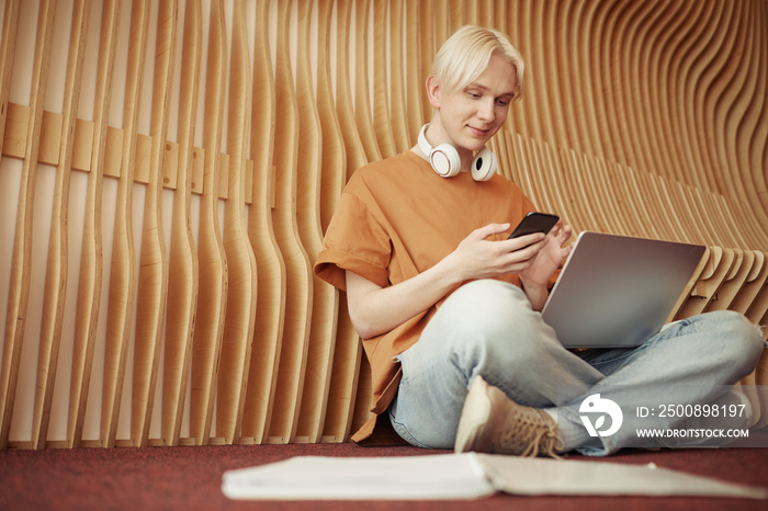 Happy blond teenage guy in casualwear texting in smartphone while sitting on the floor of college corridor at break or after classes