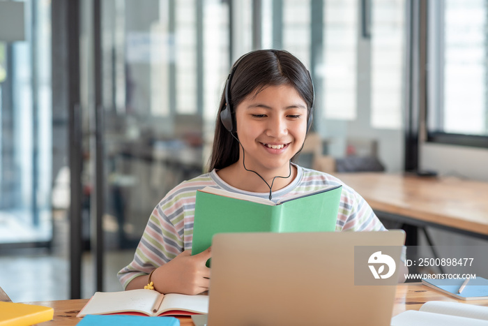 Image of a happy Asian girl wearing headphones holding a textbook online learning using a laptop in the classroom.