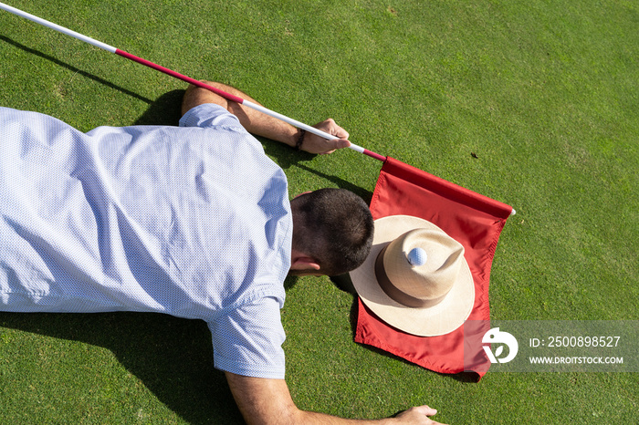 Man lying on a golf course with a hat on his side and a red flag in his hand