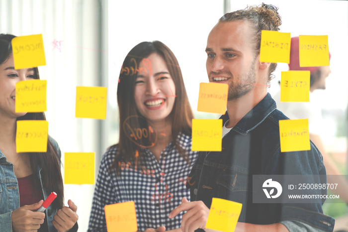 Hipster attractive young caucasian man and young asian chinese girl talk and brainstorming together at glass wall sharing business ideas project plans with post it sticky note. Marketing plan concept.