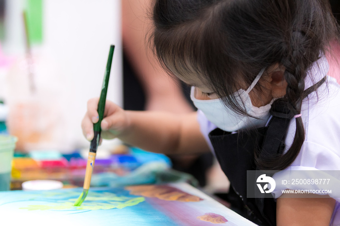 Pupils girl are concentrating on drawing and painting with brush and watercolor on the canvas. An Asian little child is wearing a white cloth face mask while learning to paint. Child 3 years old.