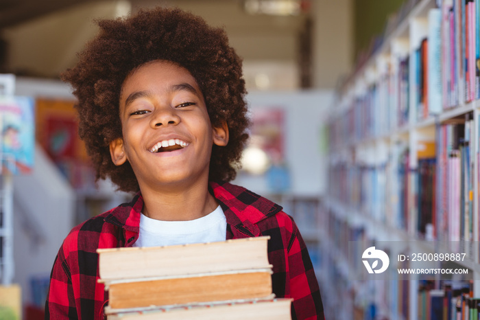 Laughing african american schoolboy carrying stack of books in school library